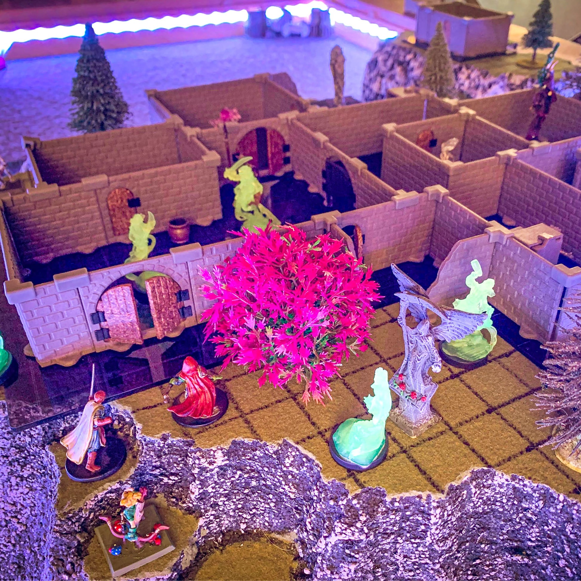 Immersive close-up of the Rogue modular dungeon walls set up on a grassy mat with fantasy terrain, featuring brightly lit pink foliage, fluorescent green "ghost" miniatures, and colorful adventurer miniatures under purple lighting.