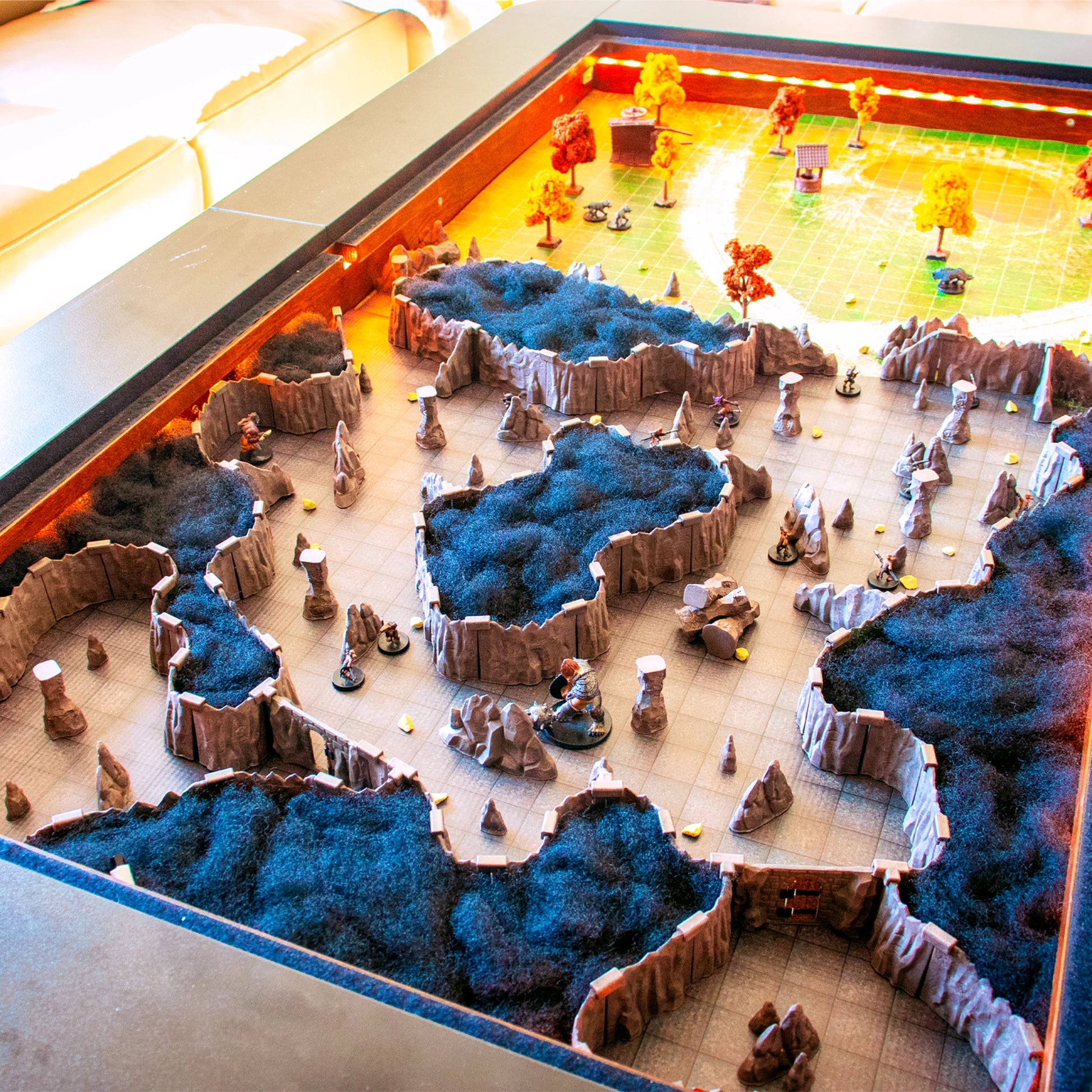 Wide view of a massive multi-level outdoor terrain setup using the Troll Cavern Set and black felt/scatter to represent elevated ground or rock. The scene includes various miniatures, trees, and scatter terrain under yellow lighting.