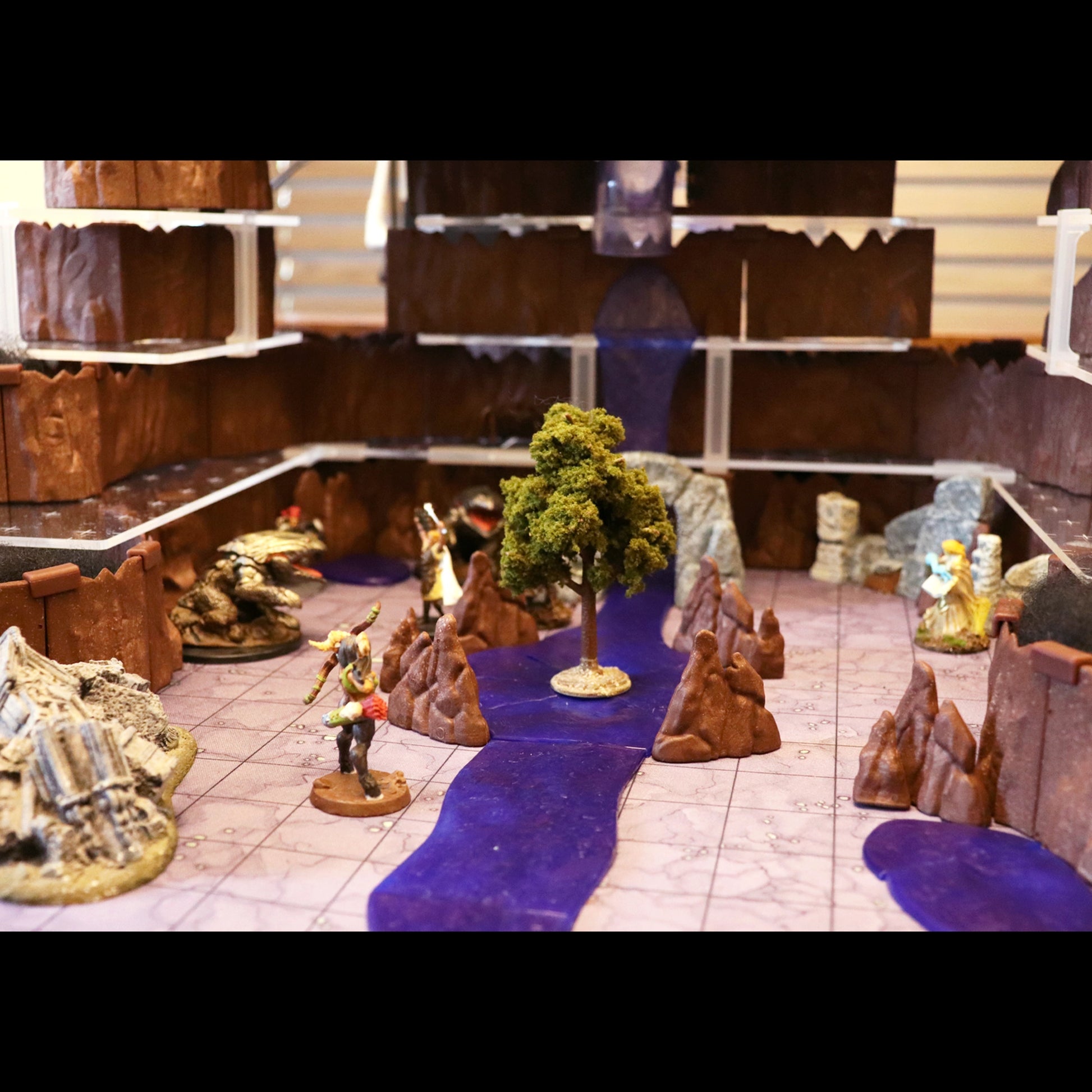 Detailed close-up of the Troll Cavern Set demonstrating terrain customization. The scene features modular brown rock pieces stacked using a Multi-Floor Expansion Combo Kit, a Waterways Kit, scattered ruins, a tree, and several fantasy miniatures.