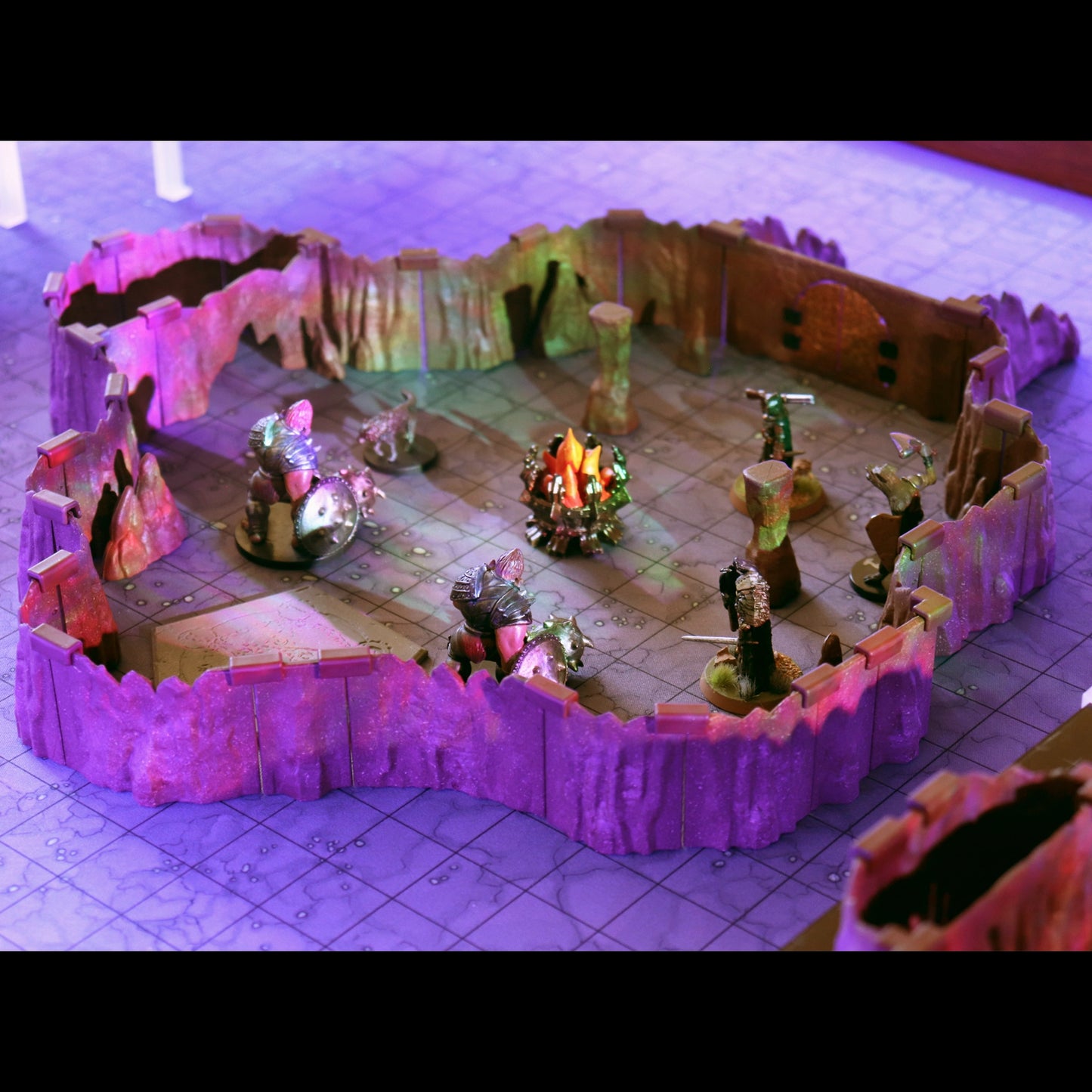 Close-up of a small, contained combat area built with the Troll Cavern walls. The scene includes multiple miniatures and a glowing campfire scatter piece, illuminated by dramatic purple ambient light.