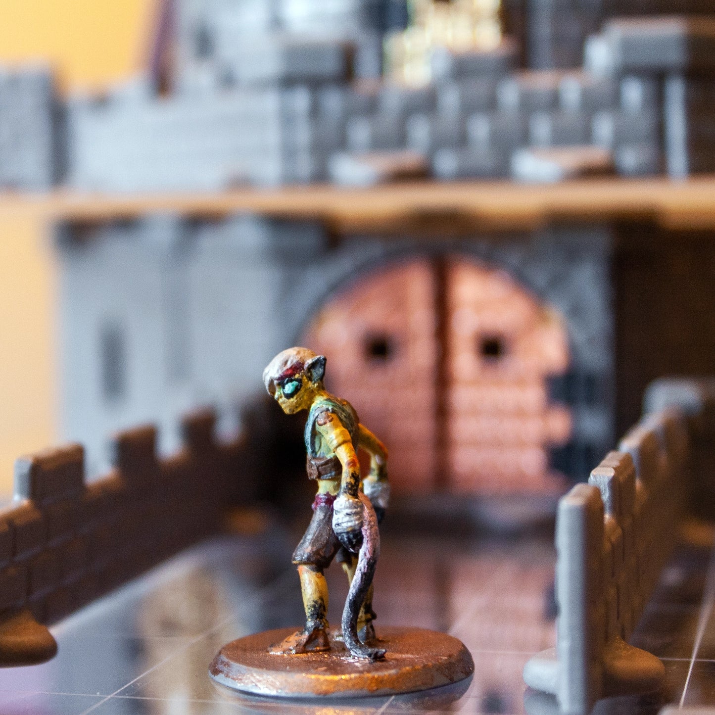 Close-up of a goblin miniature on a bridge with a large gray castle, featuring a prominent bronze metallic gate door in the background.