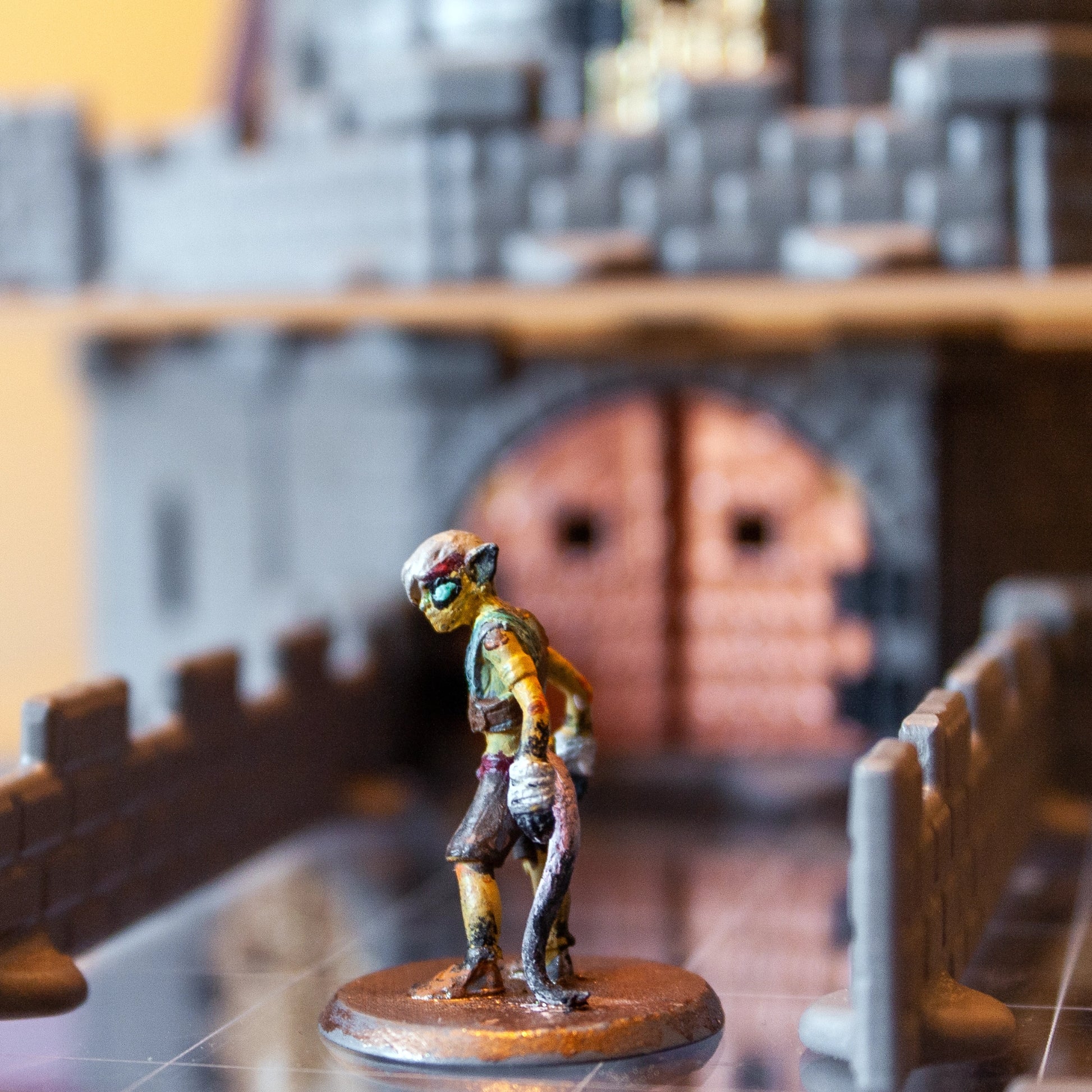 Close-up of a goblin miniature on a bridge with a large gray castle, featuring a prominent bronze metallic gate door in the background.