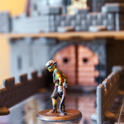 Close-up of a goblin miniature on a bridge with a large gray castle, featuring a prominent bronze metallic gate door in the background.