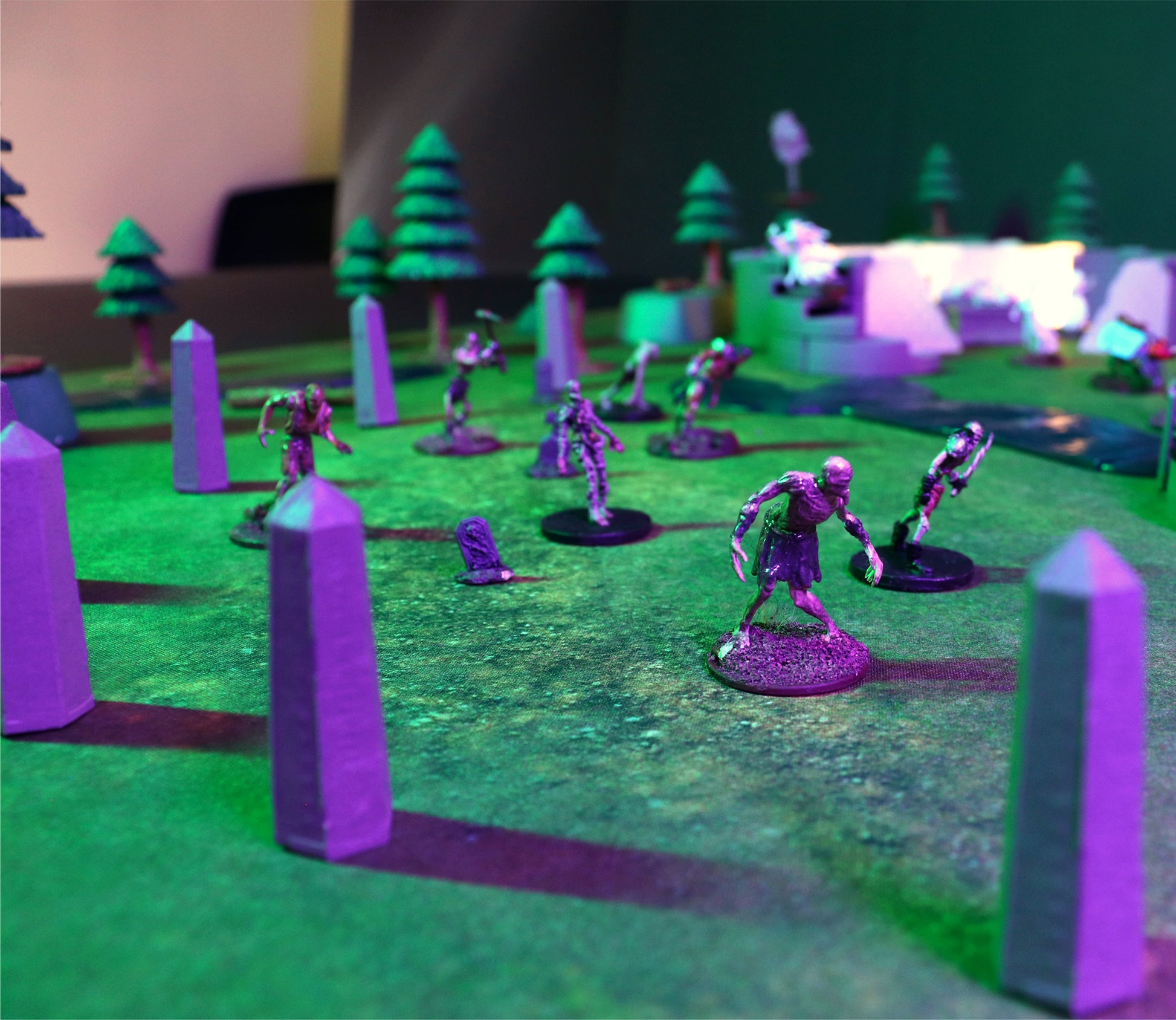 Low-angle shot of purple-lit Obelisks and a gravestone creating a cemetery terrain scene, with a horde of zombie miniatures advancing across the grass mat.