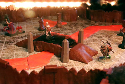Tabletop gaming scene showing a miniature standing on a central rock bridge platform over a lava flow, framed by dark dungeon and cavern walls.