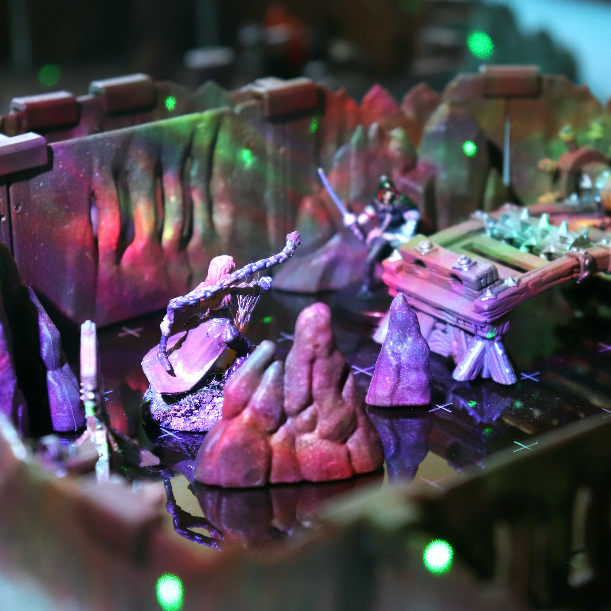Close-up of a dramatic, futuristic TTRPG scene with colorful lighting, showcasing the cavern walls, rock scatter terrain, and battling miniatures on a reflective surface.