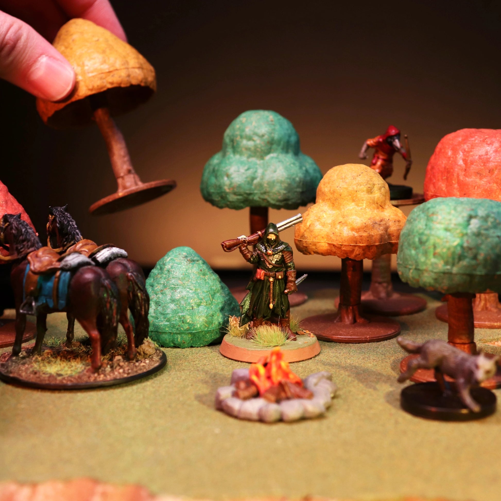 Close-up scene of a miniature in an autumn-hued forest battle, with a hand demonstrating the scale and removable canopy feature.