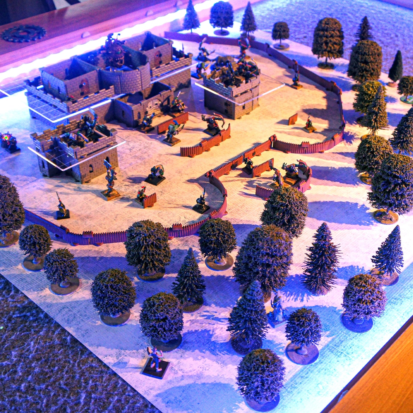 Wide view of a Baron Castle build set up as an outdoor forest fort on a green battle-mat, featuring trees, scatter terrain, and miniatures. This demonstrates the modular walls can be used for exterior scenes.