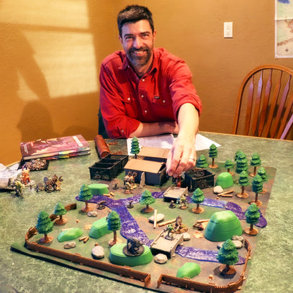 A Dungeon Master placing a miniature on a large forest terrain layout featuring the blue Waterways river sections, trees, and small buildings.