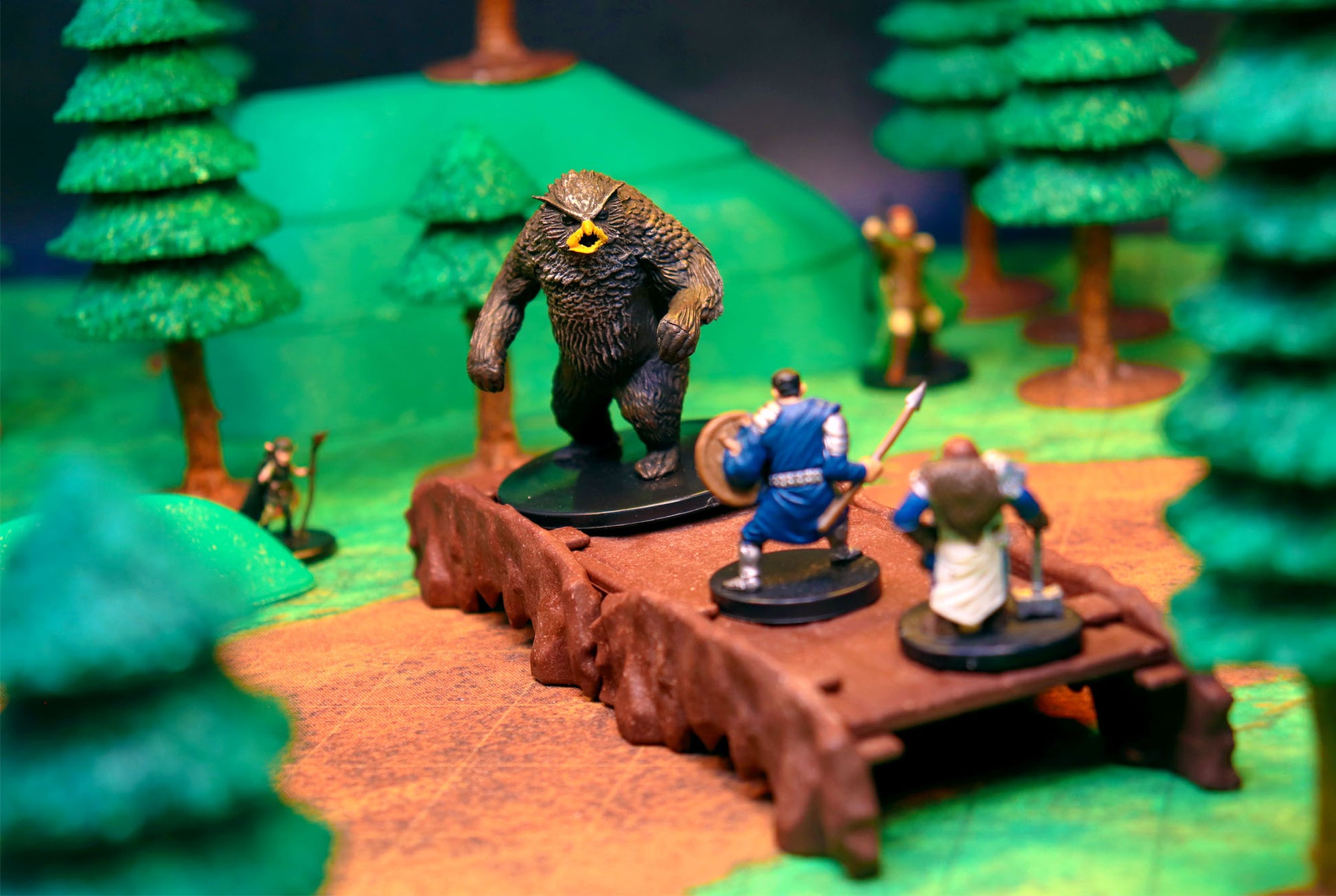 Hero miniatures fighting a large owlbear monster on a raised rock platform surrounded by the Dark Pine Forest trees.