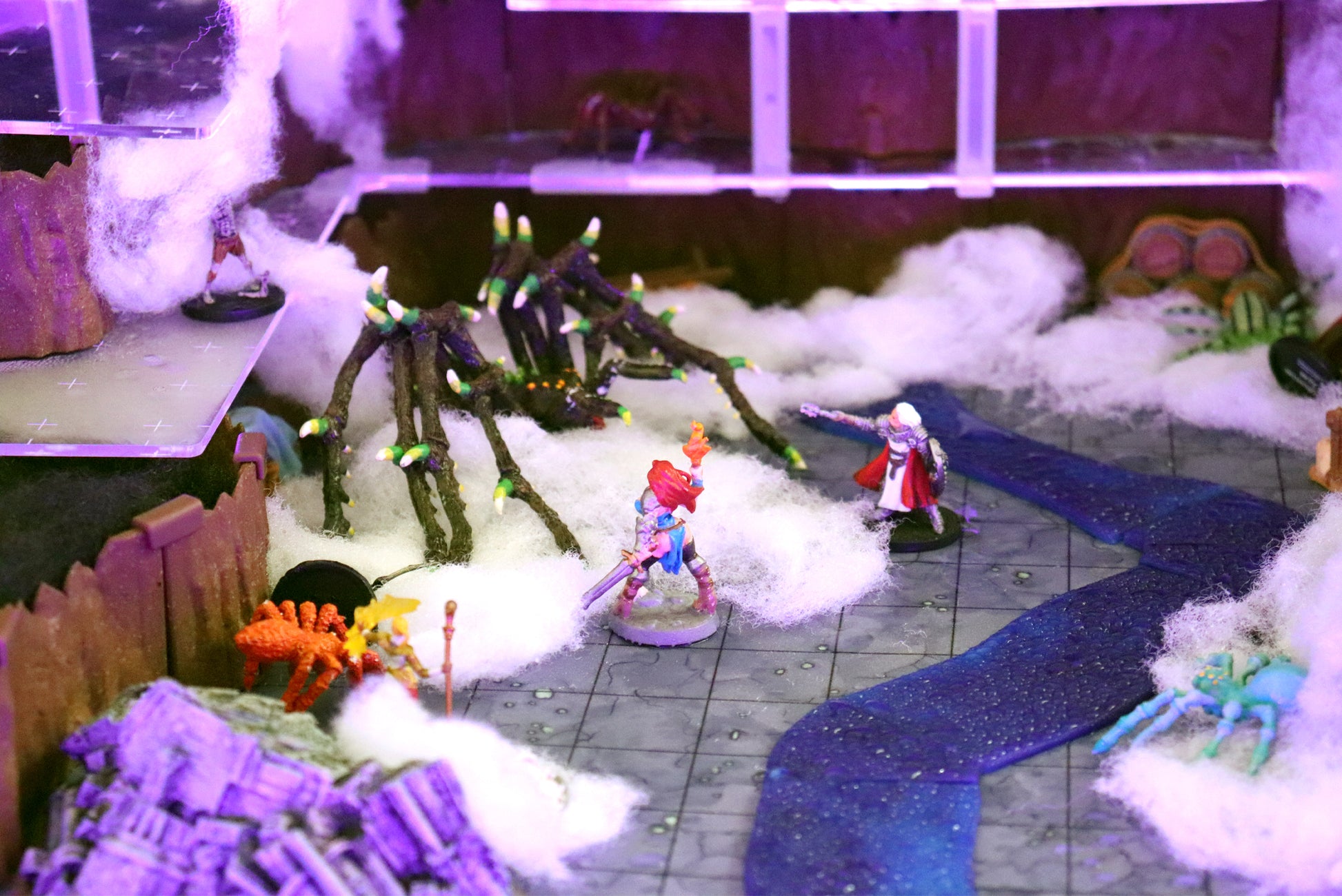 A fantasy battle scene set on a frozen river or icy terrain, featuring miniatures, cotton snow, and spider miniatures near a brown gate.