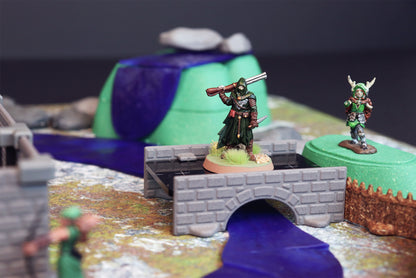 A hero miniature standing on a stone bridge accessory over a blue waterway section with a green hill in the background.