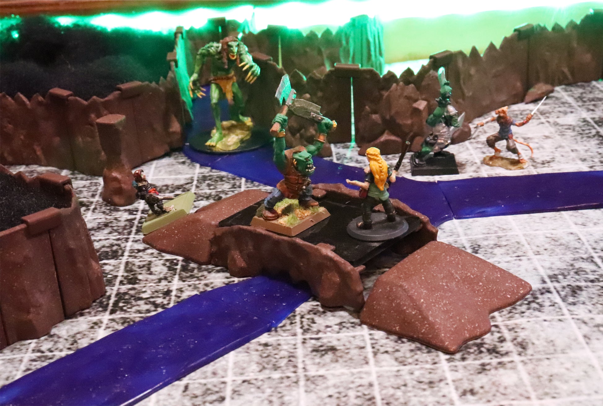 Miniatures battling near a bridge over the waterway, including a large monster on a brown rock platform, demonstrating a forest and cave setting.