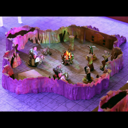 Close-up of a small, contained combat area built with the Troll Cavern walls. The scene includes multiple miniatures and a glowing campfire scatter piece, illuminated by dramatic purple ambient light.