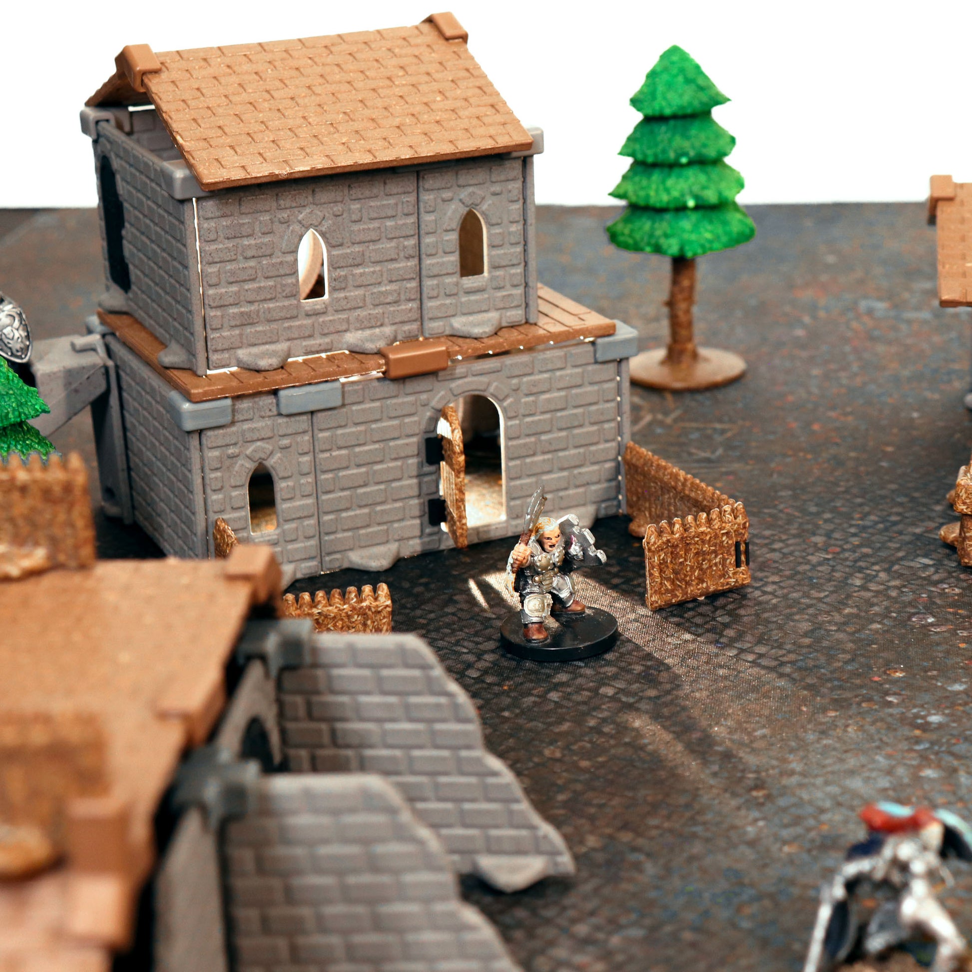 Close-up of a two-story village building with a removable brown roof, standing over a warrior miniature on a cobblestone map.